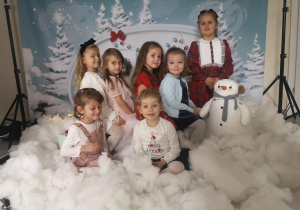 Siedem dziewczynek pozuje do zdjęcia z bałwankiem, w zimowo- świątecznej scenerii.
